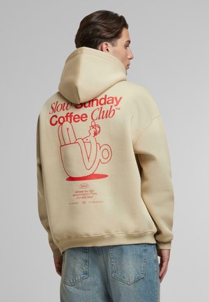 Beige hoodie featuring red graphics on the back, depicting a coffee cup and a relaxed figure. Soft fabric, loose design with a hood.