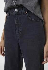 Lähikuva henkilöstä, jolla on päällään mustat korkeavyötäröiset farkut, joissa on napit ja etutaskut, sekä harmaan paidan osa ja kädet.