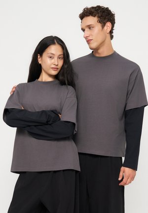 Due giovani adulti indossano maglie grigie a maniche corte abbinate sopra camicie nere a maniche lunghe e stanno vicini su uno sfondo chiaro e uniforme.