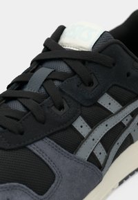 Svarta och grå sneakers har en överdel i mesh, accenter i mocka och en vadderad sula. Logotypen syns i ljusblått på tungan.