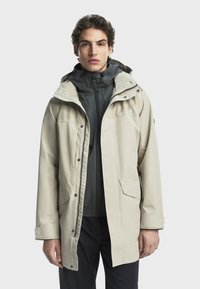 Svijetlosmeđi vodootporni jakna s kapuljačom, koja ima zatvarač i kopčanje na dugmad, podesive manšete i prednje džepove. Preko tamne unutarnje odjeće.