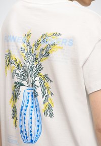 Tricou alb din bumbac, cu un imprimeu pe spate reprezentând un vas floral albastru și galben, cu detalii text în albastru deschis și verde închis.