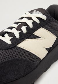 Baskets noires avec une tige en maille et des accents en daim, arborant un logo "N" couleur crème, des lacets blancs et une semelle en caoutchouc texturée.