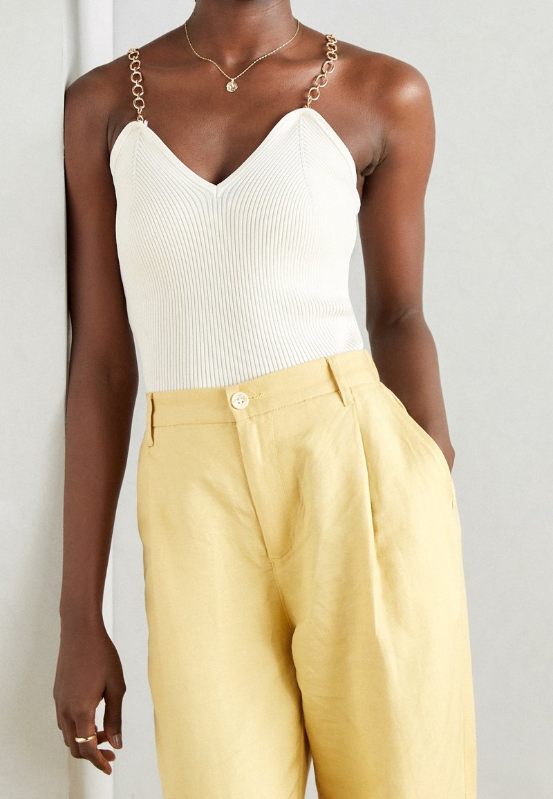 White ribbed tank top with V-neck and gold chain straps, paired with loose yellow linen trousers featuring a front button closure.