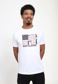Bílý bavlněný tričko s opotřebovanou grafikou loga MTV a červenými a modrými pruhy připomínajícími americkou vlajku. Krátké rukávy, kulatý výstřih.