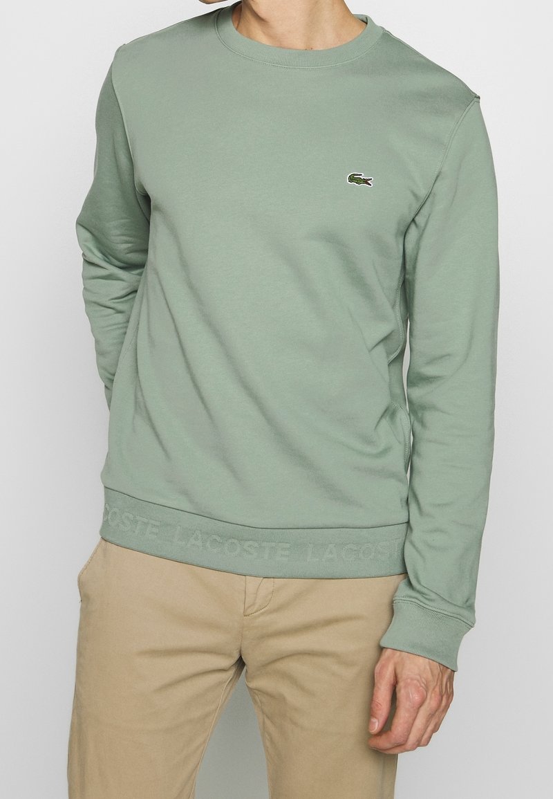 Homme portant un sweat-shirt vert clair à col rond avec un petit logo crocodile sur la poitrine gauche et un pantalon beige, debout avec une main derrière le dos.