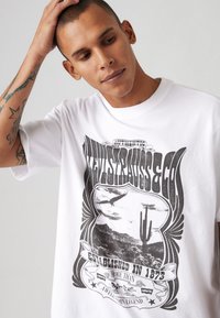 Balta medvilninė marškinėlių su vintage stiliaus juodu grafiniu piešiniu, vaizduojančiu dykumos sceną su kaktusu ir ereliu. Tekstas skelbia "Levi's Strauss & Co."