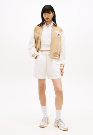 Fiatal nő áll, bézs és fehér varsity dzsekit, fehér kötött rövid pulóvert, fehér rövidnadrágot, fehér zoknit és ezüst tornacipőt visel.