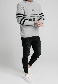 Light gray knit sweater with black stripes and logo, paired with ripped black skinny jeans and white sneakers. Accents include textured fabric.