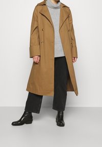 Brauner Trenchcoat mit großen Revers und doppelter Knopfleiste, getragen über einem grauen Rollkragenpullover und schwarzen Jeans; kombiniert mit schwarzen Knöchelstiefeln.