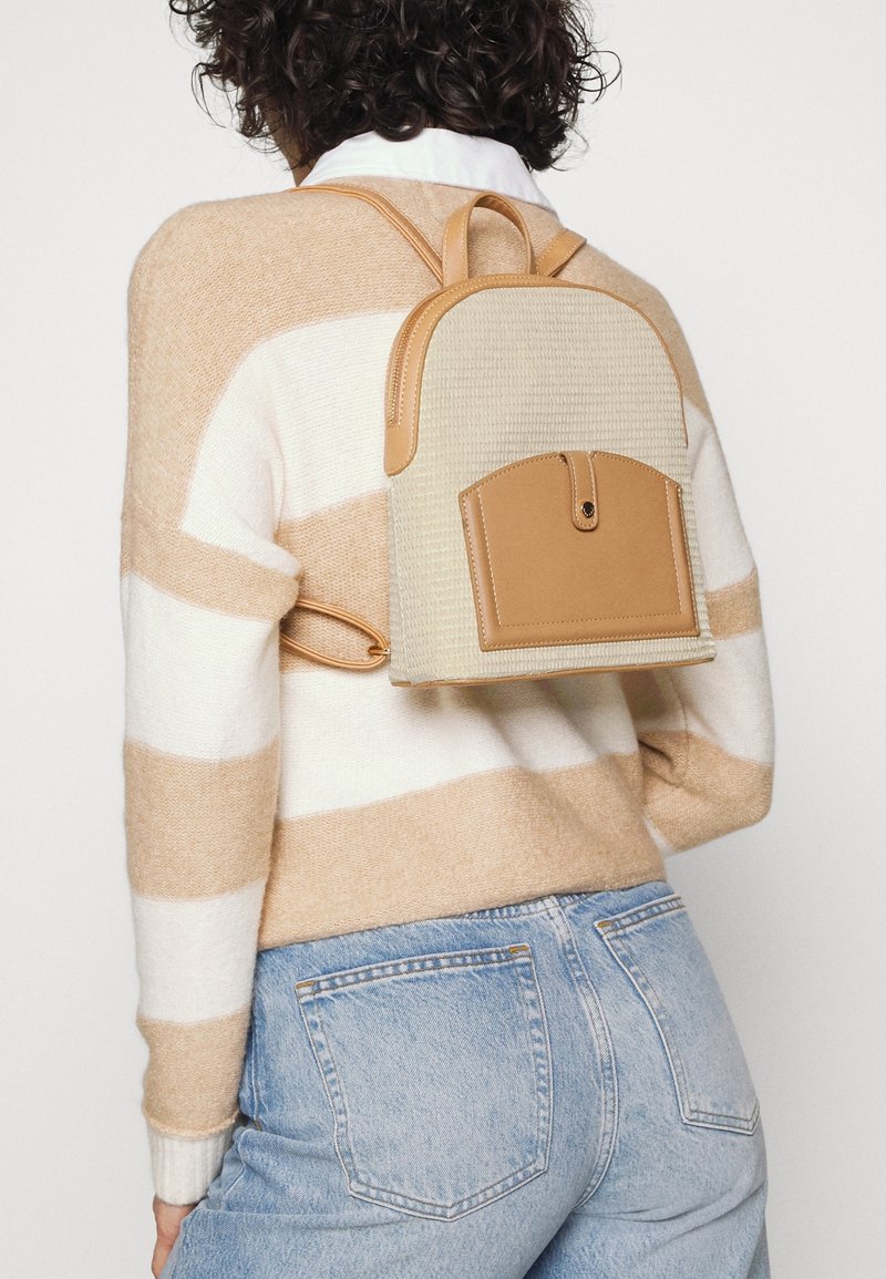 Personne portant un pull rayé beige et blanc et un jean bleu, portant un petit sac à dos beige et marron clair avec poche avant.
