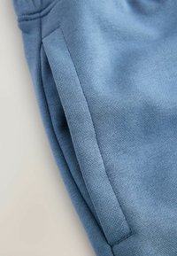 Gros plan d'un vêtement en tissu bleu montrant une poche latérale avec des coutures visibles et un matériau tricoté texturé.