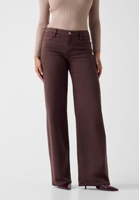 Jeans marroni a gamba larga con vita alta, chiusura con bottone metallico e design a cinque tasche, abbinati a un top beige a maniche lunghe con costine.