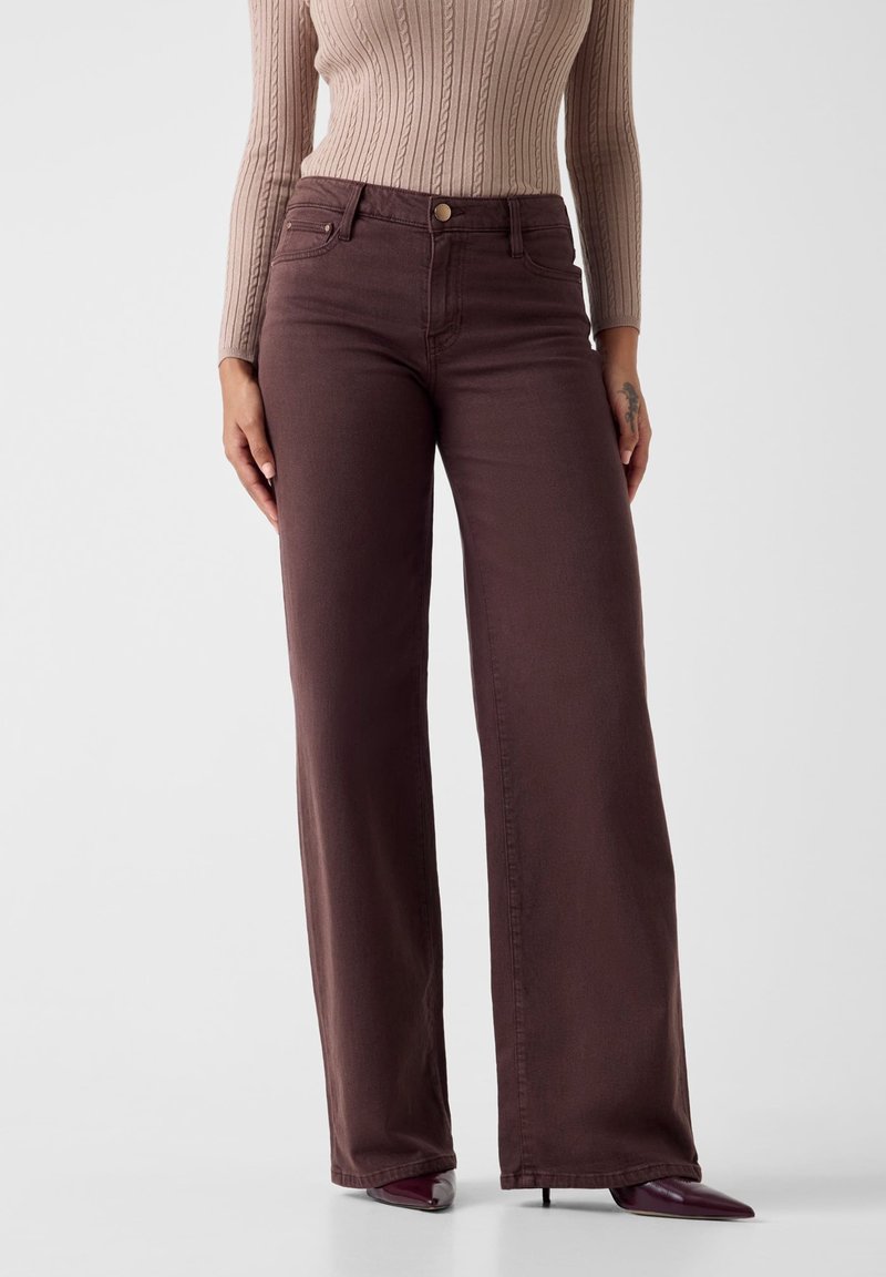 Jeans marroni a gamba larga con vita alta, chiusura con bottone metallico e design a cinque tasche, abbinati a un top beige a maniche lunghe con costine.