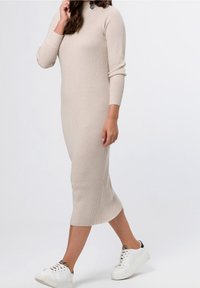 Abito a maglia beige a coste con collo alto e maniche lunghe, caratterizzato da una silhouette leggermente aderente e uno spacco laterale. Abbinato a sneaker bianche.