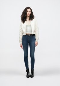 Κοντό κρεμ μπουφάν bomber πάνω από γκρι αμάνικη μπλούζα, συνδυασμένο με στενά σκούρα μπλε τζιν και μαύρα μποτάκια στον αστράγαλο, με ασημένια ζώνη.