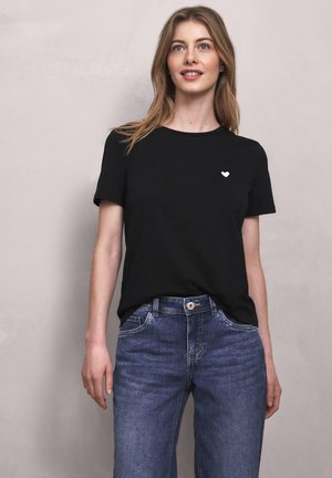 Mladá žena s dlouhými vlasy, která má na sobě černé tričko s malým bílým srdcem na hrudi a modré džíny, mírně se usmívá a stojí.