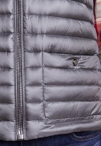 Šedá péřová vesta vyrobená z lehkého, lesklého materiálu s prošívanou strukturou, přední zip a drukový detail na hrudi.