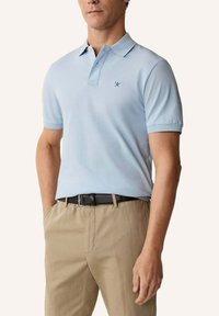 Homme portant un polo à manches courtes bleu clair avec un logo de rames croisées, un pantalon beige et une ceinture noire, debout avec une main dans la poche.