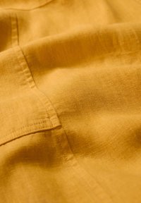 Žlutá lněná látka s hladkou texturou, zdobená jemným prošíváním a záhyby, které zdůrazňují měkký a lehký vzhled.