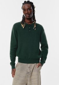 Tmavohnedý pulóver so stojacím golierom, trojiciou gombíkov a rebrovanými manžetami. Nosený s svetlými džínsami.