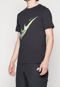 Černé bavlněné tričko s velkým stylizovaným logem Nike v šedé a neonově žluté barvě na přední straně. Krátké rukávy a kulatý výstřih.