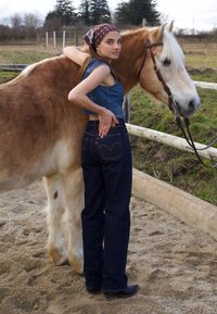 Moteris deniminiu trumpu viršumi ir tamsiomis džinsomis stovi šalia šviesiai rudo arklio, viena ranka laikydama arkliui kaklą.