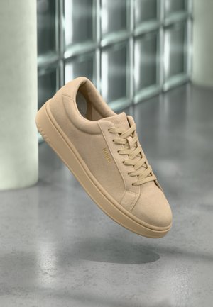 Beige suede low-top sneaker with laces and "HUGO" logo on side, set against a reflective glass block wall and gray floor background.
