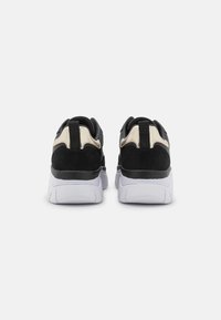 Un paio di sneakers nere e beige con suole bianche spesse, mostrate da dietro su uno sfondo grigio chiaro uniforme.