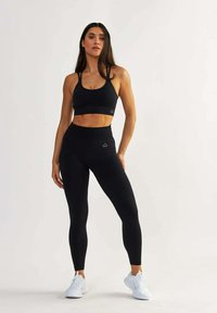 Schwarzer Sport-BH und hochgeschnittene Leggings aus strukturiertem, stretchigem Material. Weiße Logo-Akzente und kombiniert mit weißen Sportschuhen.