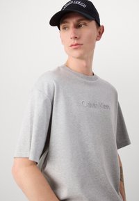 Light gray short-sleeve T-shirt featuring an embossed Calvin Klein logo. Smooth cotton fabric with a relaxed fit and crew neckline.