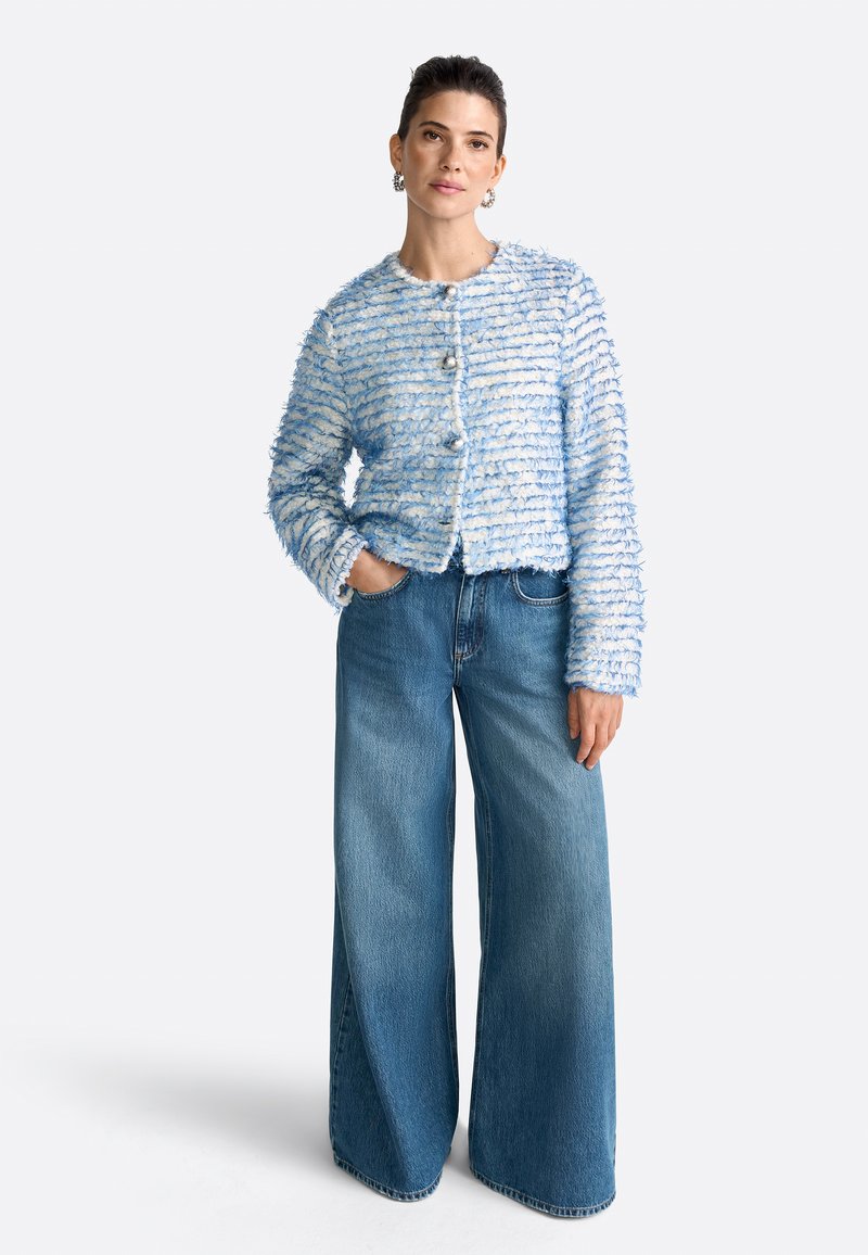 Ljusblå texturerad jacka med vita ränder, med rund halsringning, knappstängning framtill och långa ärmar. Kombinerad med vida blå jeans.