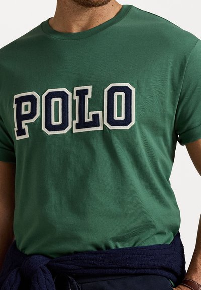 T-shirt verde de manga curta feita de algodão, apresentando grandes letras brancas e azuis-marinho a soletrar "POLO" na parte da frente. Textura suave, design de gola redonda.