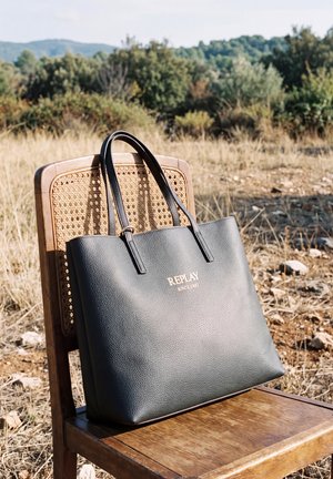 Un sac cabas en cuir noir repose sur une chaise en bois à dossier en cannage, dans un champ ensoleillé avec de l’herbe sèche et des arbustes.