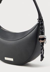 Fekete félhold alakú bőrtáska ezüst színű márkajelzéssel és fekete-fehér zsinórral, amelyen egy napszemüveges figura mesefigura bábu lóg.