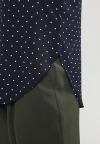 Námořnická modrá halenka s puntíky a zakřiveným lemem, doplněná olivově zelenými kalhotami. Halenka má bílé puntíky a hladkou texturu látky.