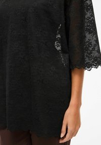 Czarna koronkowa bluzka z luźnymi rękawami długości do łokcia, z fakturowanym wzorem w kratę i falistym brzegiem. Ręka spoczywa na materiale.