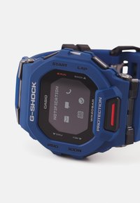 G-SHOCK digital klocka med ett blått silikonsnöre, svart display och röd accentknapp. Funktioner inkluderar notifieringsikoner och vattentålighet.