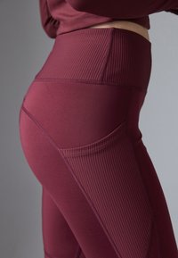 Λήψη κοντινού πλάνου της μέσης και του γοφού ενός ατόμου που φοράει μπορντό ψηλόμεσα κολάν με ραβδωτή πλευρική τσέπη και ταιριαστό crop top.