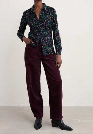 Persona con camisa oscura de manga larga con estampado floral, pantalones burdeos de pierna ancha de talle alto y botines negros puntiagudos, de pie con una mano en el bolsillo.