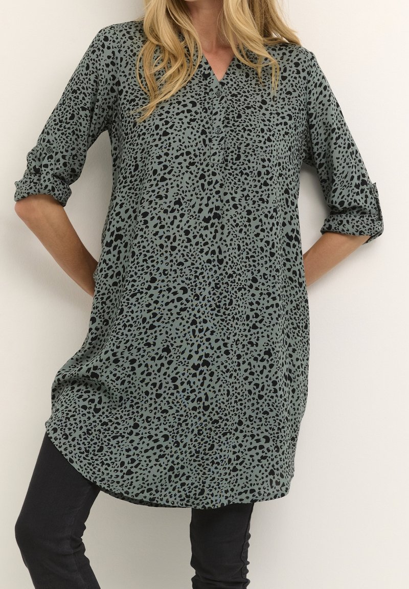 Femme portant une longue tunique verte à taches léopard noires avec manches retroussées, associée à un pantalon noir ajusté.
