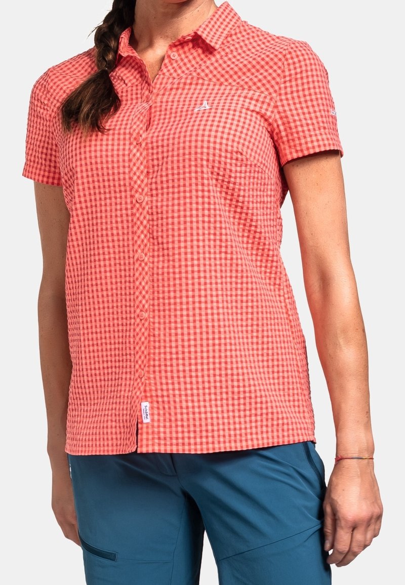 Femme portant une chemise boutonnée à manches courtes à carreaux corail et rouge et un pantalon bleu, debout avec sa main gauche détendue le long du corps.