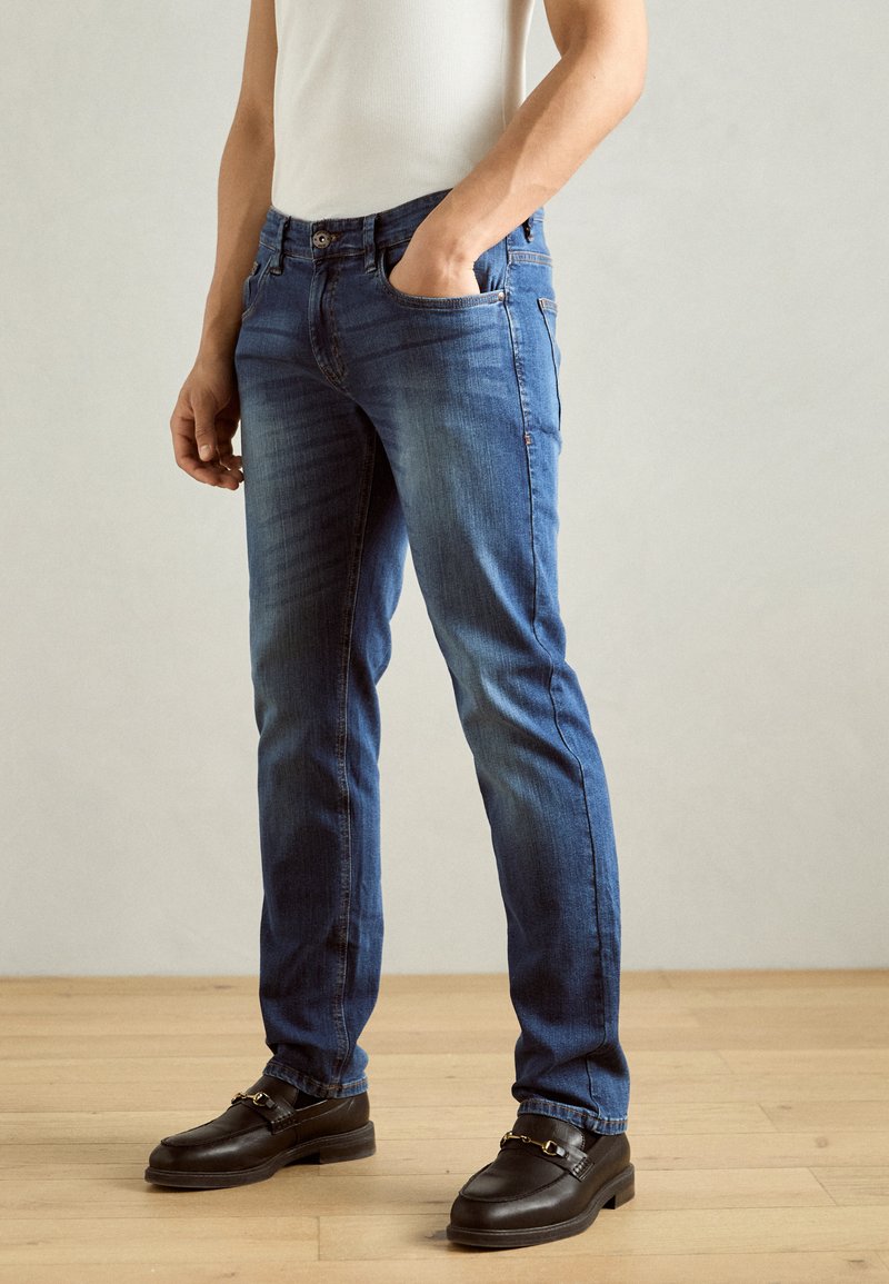Denim jeans i en middels blå vask med slim fit, med en frontlomme, beltehemper og subtile falmende detaljer. Kombinert med brune loafers.