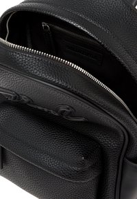 Črn usnjen nahrbtnik z strukturirano površino, zadrgo, notranjim žepom in vidno oznako logotipa, ki prikazuje eleganten, sodoben dizajn.