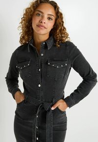 Vestido de camisa de mezclilla negra con mangas largas, dos bolsillos en el pecho con tachuelas plateadas, cintura ceñida con cinturón y frente abotonado. Textura suave.