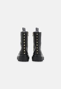Botas negras hasta el tobillo con un acabado brillante, tachuelas doradas a los lados, suelas gruesas y tiradores con una marca blanca en la parte superior.