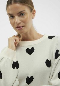 Pull léger beige avec motifs de cœurs noirs, présentant un col et des poignets côtelés, et une coupe ample avec un tissu texturé.