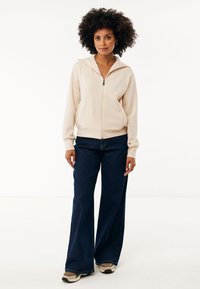 Sudadera cremosa con cremallera y capucha, acompañada de jeans de pierna ancha de color azul oscuro y zapatillas beige. Diseño sencillo, textura suave.