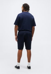 Άνδρας με πλεξούδες, που φοράει μπλε ναυτικό T-shirt και σορτς, άσπρες κάλτσες και μαύρα αθλητικά παπούτσια, στέκεται με την πλάτη προς το μέρος μας σε απλό φόντο.