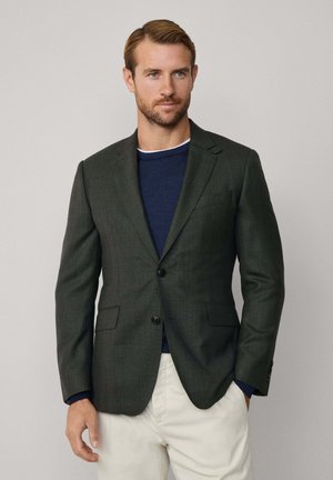 Dunkelgrünes Blazer aus strukturiertem Stoff, einreihiger Schnitt, zwei Knöpfe und Reverskragen, kombiniert mit einem navyblauen Sweater und cremefarbener Hose.